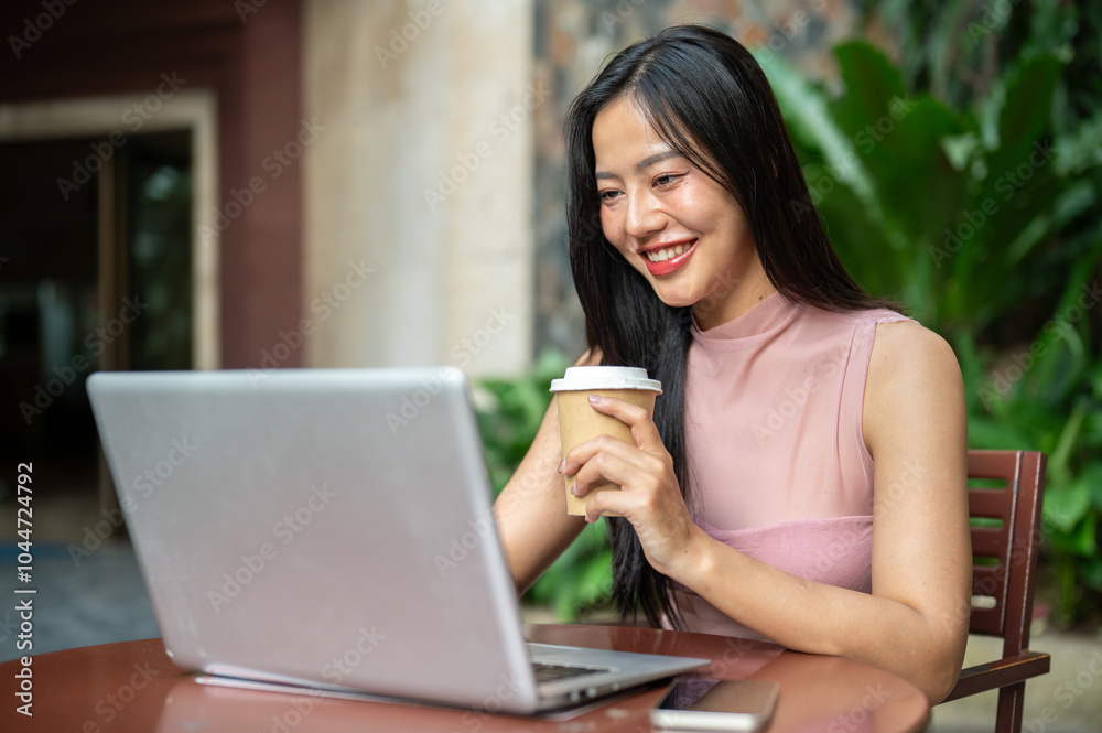 A happy woman is working remotely from a coffee shop, enjoying coffee while working on her laptop. A happy woman is working remotely from a coffee shop, enjoying coffee while working on her laptop.