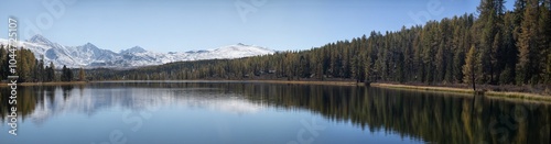 Panoramic landscape of a large mountain lake in a northern nature: snowy mountain peaks, dense coniferous forest. The season is golden autumn, September: yellow and red red grass, golden foliage.