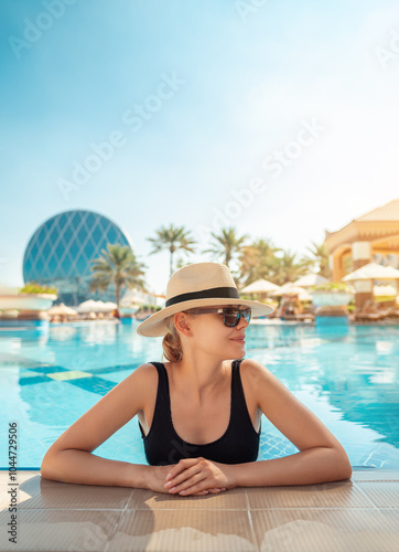 Girl in hat is in pool, leaning on side. Young woman with glasses and hat on vacation is relaxing on edge of pool on sunny day. Portrait of woman in pool looking at camera. Sitting on edge of pool