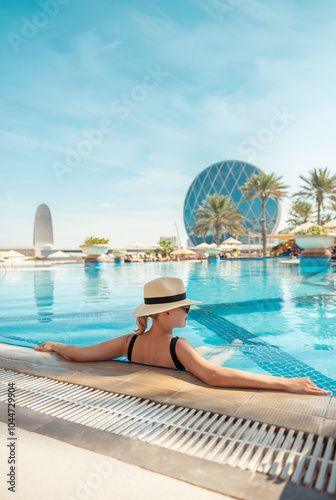 Girl in hat is in pool, leaning on side. Young woman with glasses and hat on vacation is relaxing on edge of pool on sunny day. Portrait of woman in pool looking at camera. Sitting on edge of pool