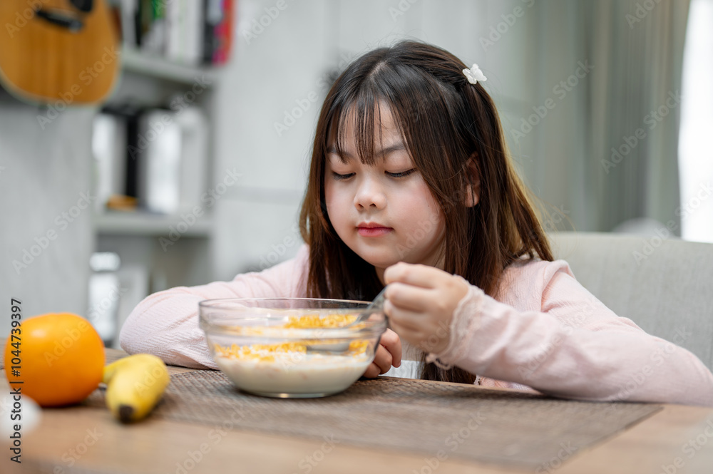 A young, adorable Asian girl is eating a bowl of cereal at the dining table for breakfast. A young, adorable Asian girl is eating a bowl of cereal at the dining table for breakfast.