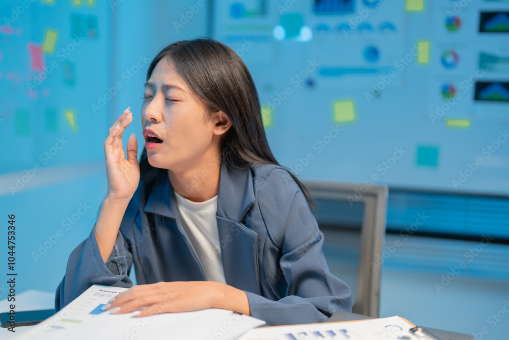 Tired businesswoman yawns at her desk in a dimly lit office, surrounded by paperwork, feeling exhausted and stressed from working late