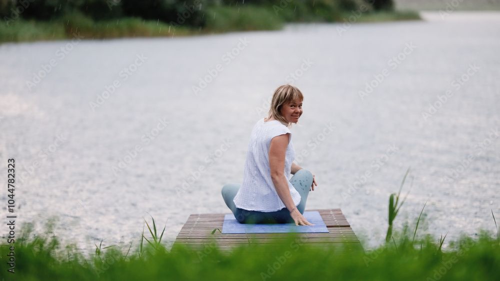 Fototapeta premium Young Caucasian woman sitting in lotus position on bridge by lake during rain. Woman enjoying fresh air. Outdoor meditation