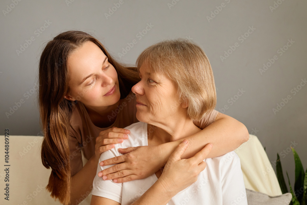 Elderly Caucasian woman and her adult daughter sitting together on couch smiling sharing joyful moment together at home hugging with love and looking at each other Elderly Caucasian woman and her adult daughter sitting together on couch smiling sharing joyful moment together at home hugging with love and looking at each other