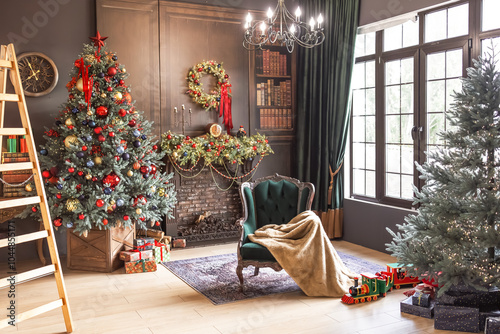 warm living space decorated holiday cozy stylish designer living room interior. Two Christmas trees are adorned with colorful ornaments and lights. green armchair draped soft blanket sits nearby.