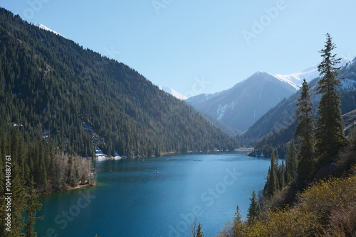 Pristine mountain lake surrounded by evergreen forest and snow-capped peaks, turquoise waters reflecting autumn colors. Kolsai lake, Kazakhstan.