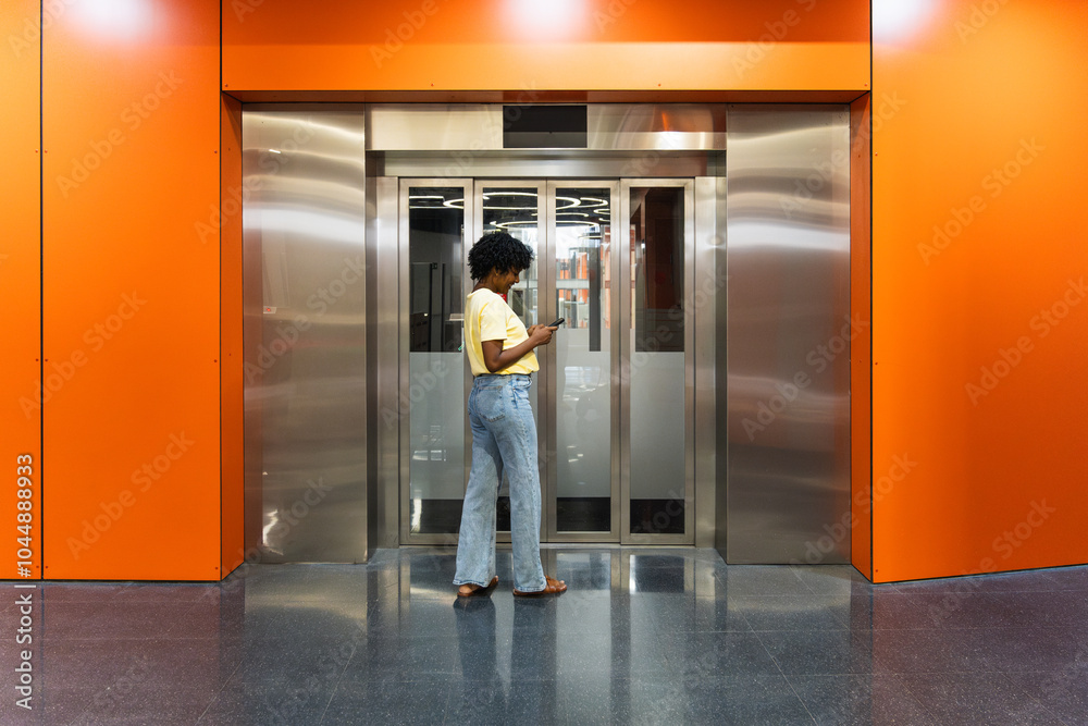Cheerful African American woman using smartphone while waiting for elevator in hotel lobby Cheerful African American woman using smartphone while waiting for elevator in hotel lobby
