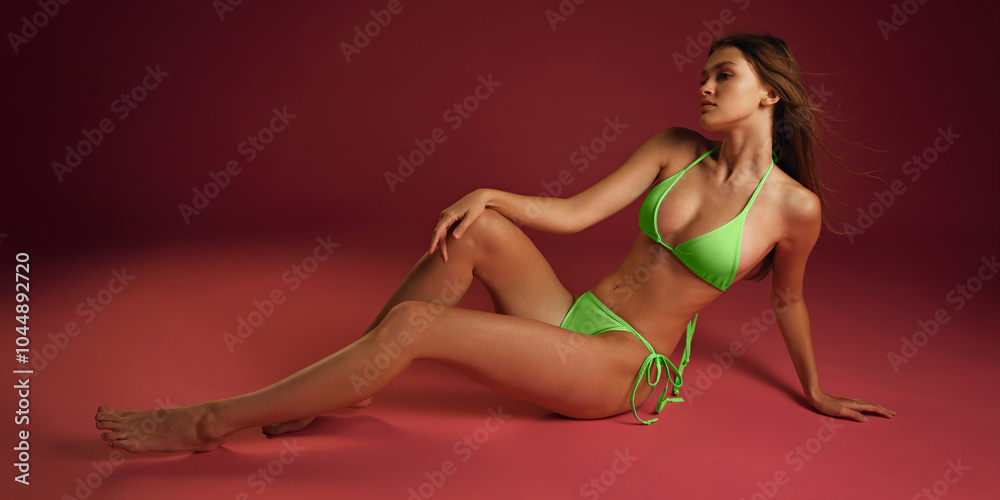 Fototapeta premium Full-body shot of woman posing, lying confidently in vibrant green bikini against burgundy color background. Relax mood. Concept of female health dieting, natural beauty, wellness. Ad