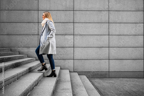 Middle-aged blonde woman in gray coat, scarf, jeans and sunglasses walking up concrete stairs  against background of concrete slabs in city on autumn day. Businesswoman. Side view.