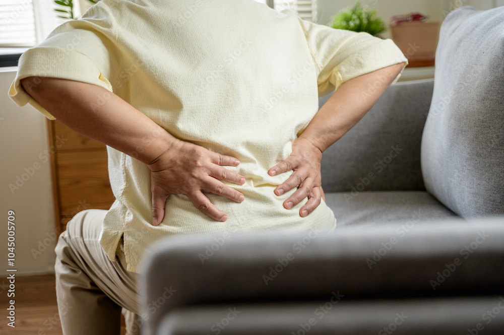Fototapeta premium Senior Asian woman using hand pushing on her back because feeling back pain. An elderly person is sitting on a couch, holding their lower back in pain. highlighting discomfort and the need for medical