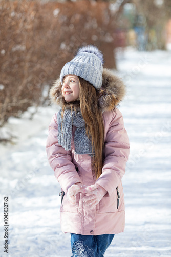 Happy little girl in warm pink coat and grey knitted hat has fun and walking in snowy winter park. Children outdoor activities concept. Happy family holidays time. High quality photo