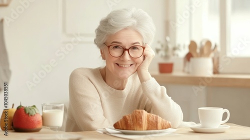 A cheerful senior woman enjoys breakfast with croissants and coffee at a cozy table, highlighting a peaceful morning and healthy lifestyle.
