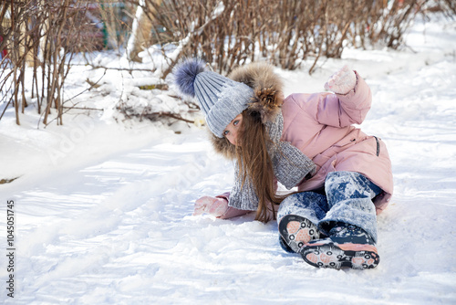 Happy excited little girl in warm clothes sitting on a snow, happily playing in winter park and joyfully laughing. Fun outdoor activities for children in wintertime. Winter portrait with copy space #1045051745