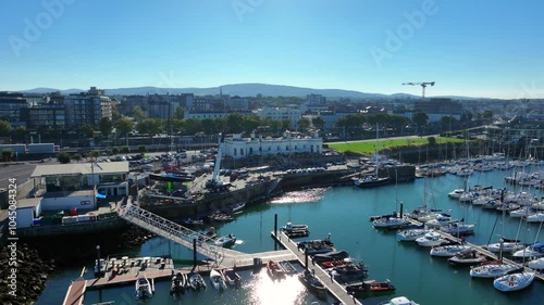 Royal Irish Yacht Club, Dún Laoghaire, County Dublin, Ireland, September 2024. Drone orbits counter clockwise as sunlight shimmers across water with yachts and sailboats moored in the Marina. photo