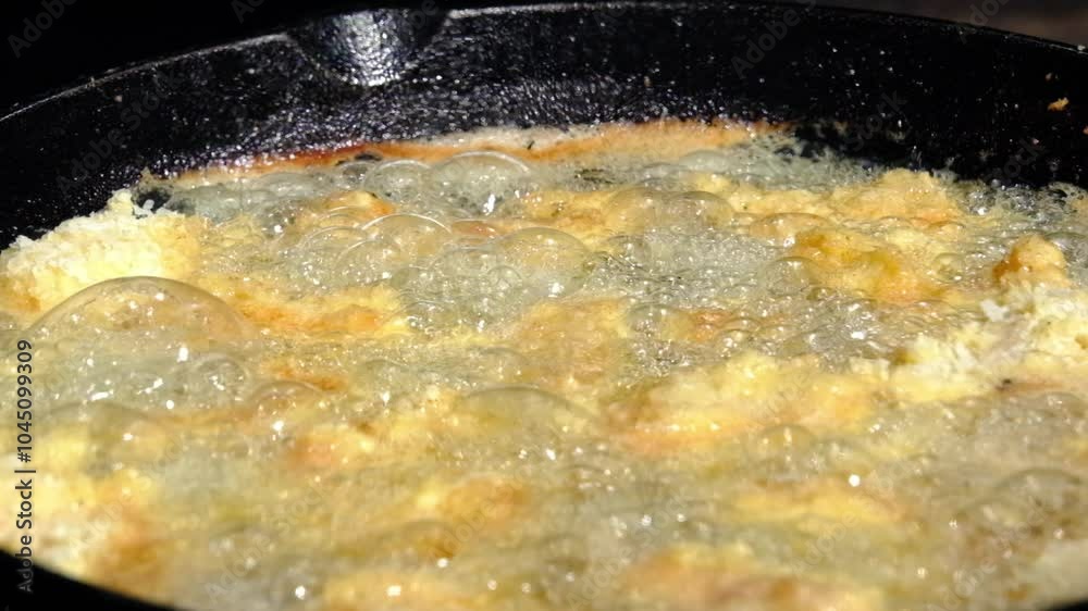 Slow motion and close up of catfish nuggets or fish battered in breadcrumbs being deep fried in cast iron skillet or pan over firepit or camp fire
