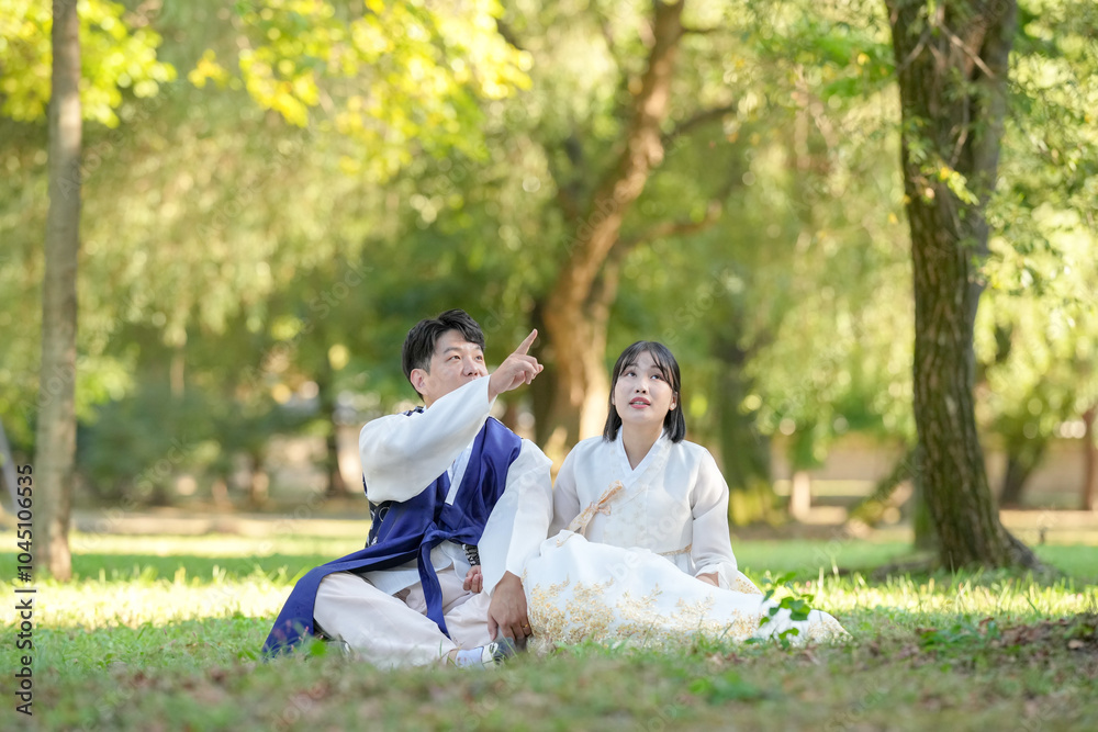 A Korean couple in their 30s and a woman in their 20s are looking at the sky in a green park surrounded by trees while wearing Hanbok in Seoul, South Korea.