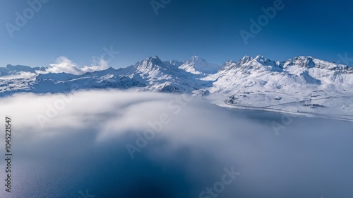 Aerial view of snow-capped mountains with fog rolling through a valley below, clear blue sky.