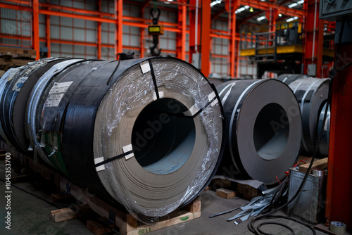 A large roll of metal is wrapped in plastic and sits on a wooden pallet. The roll is surrounded by other rolls of metal, all of which are wrapped in plastic. The scene is industrial in nature