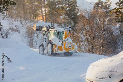 Snow plow tractor cleaning snow with a large rear-mounted snowblower, shooting snow to the side. Mountain area with snow-covered road. Effective snow removal, rural road access.