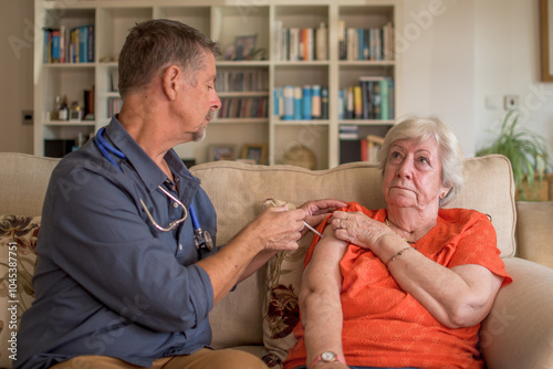 A physician, doctor injects the winter flu jab into a elderly lady at home. Covid, influenza vaccination, preventative care administered by a visiting GP. Home healthcare.