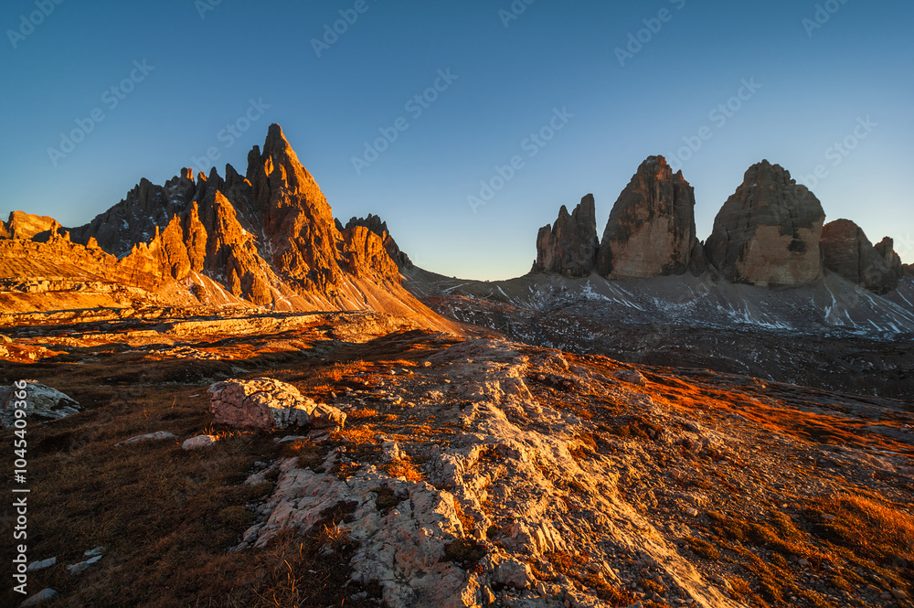 Obraz premium Majestic peaks of the Tre Cime di Lavaredo, Dolomites bathed in golden sunlight at dusk as the rugged landscape showcases nature’s beauty in Italy's renowned mountain range.