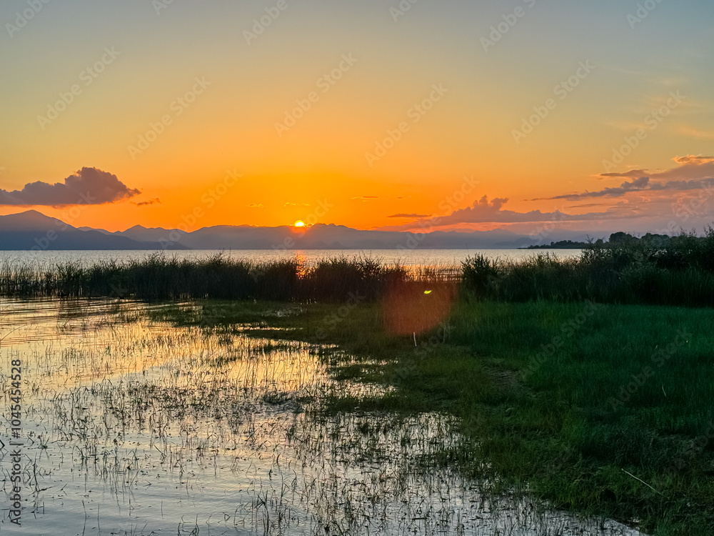 Breathtaking sunset over calm Skadar lake, Albania. Sun is setting behind Dinaric Alps mountains in Montenegro. Water reflects vibrant colors of sky creating shimmering path across the surface Breathtaking sunset over calm Skadar lake, Albania. Sun is setting behind Dinaric Alps mountains in Montenegro. Water reflects vibrant colors of sky creating shimmering path across the surface