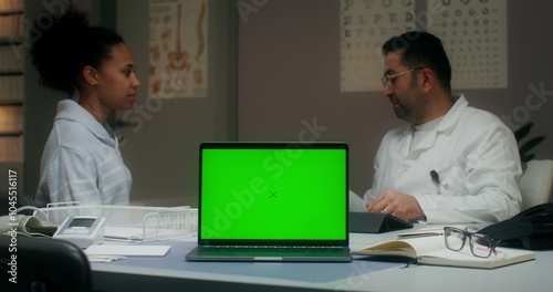 A laptop with a green screen is on the table in the medical office. The doctor measures the temperature of a female patient