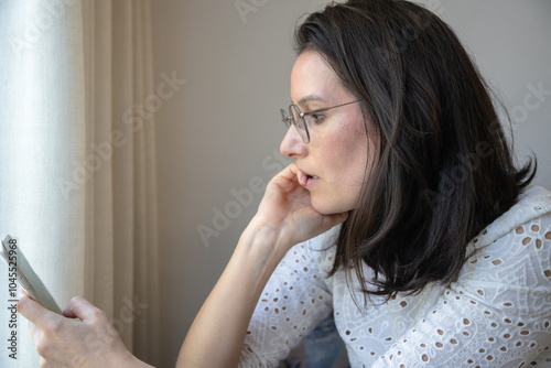 A woman in glasses looks intently at her smartphone with a thoughtful expression, sitting by a window in soft natural light. She appears focused, absorbed in reading or texting.