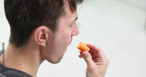 Man eats bright thin carrot chewing bit off piece with pleasure. Man enjoys taste of fresh vegetable carefully examining product in hand slow motion