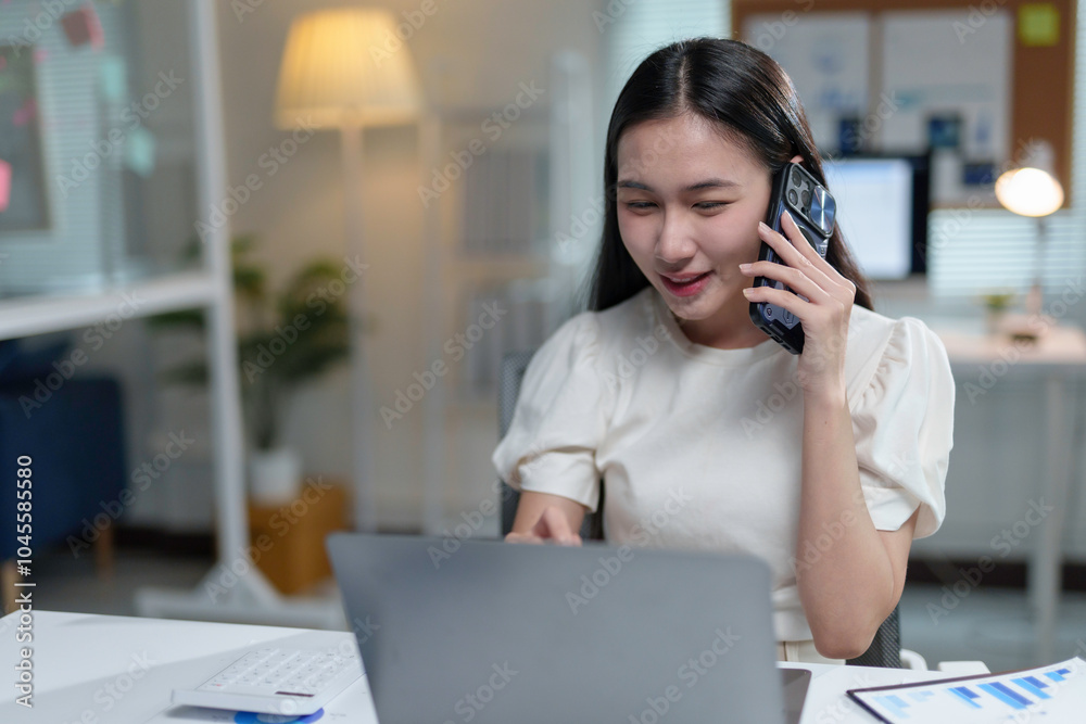 Asian businesswoman is multitasking, talking on her phone while working on her laptop in her office Asian businesswoman is multitasking, talking on her phone while working on her laptop in her office