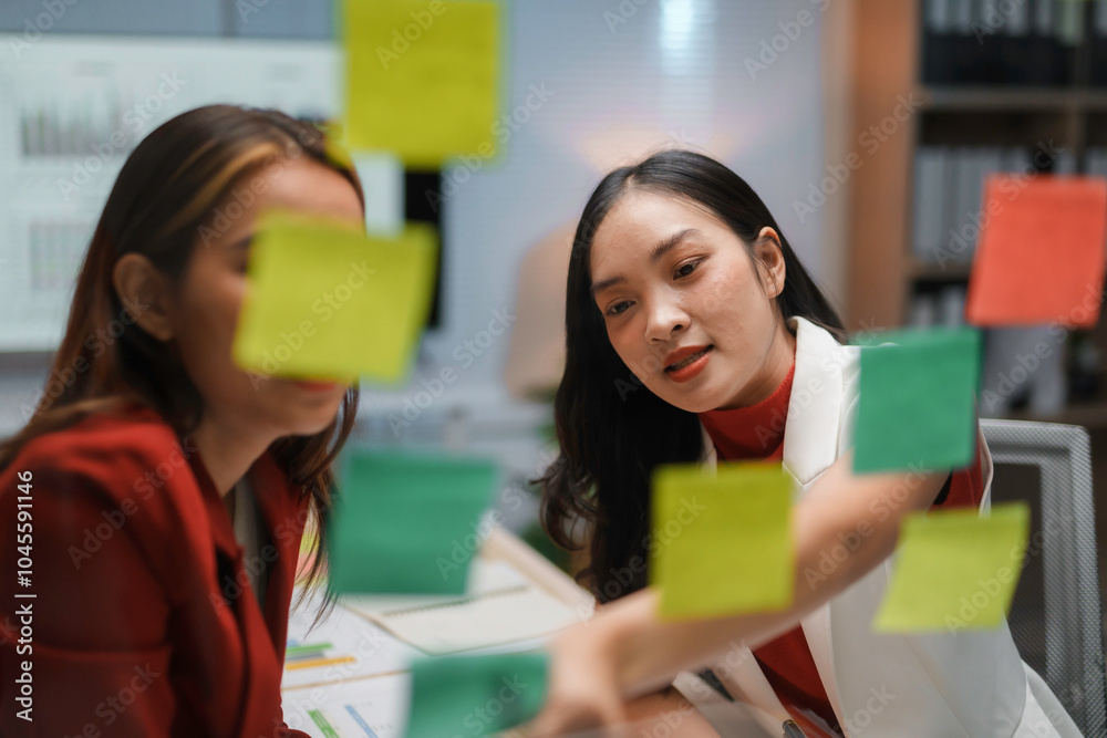 Two businesswomen discussing and planning their tasks using sticky notes on a glass wall in the office at night Two businesswomen discussing and planning their tasks using sticky notes on a glass wall in the office at night