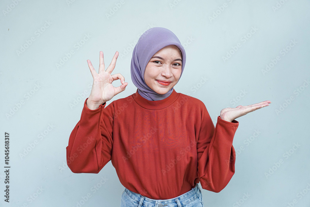 Beautiful smiling Asian woman in red sweater showing okay and pointing or showing gesture demonstrates symbol of approval isolated over gray background.
