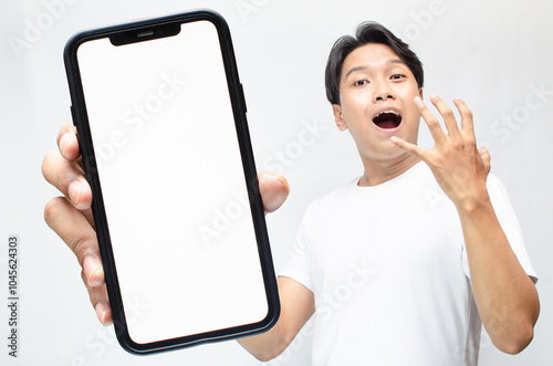 mock up of young Asian man showing blank screen smartphone while making yes gesture.  asian man in white tshirt holding and showing phone screen while celebrating something and raising his fist.