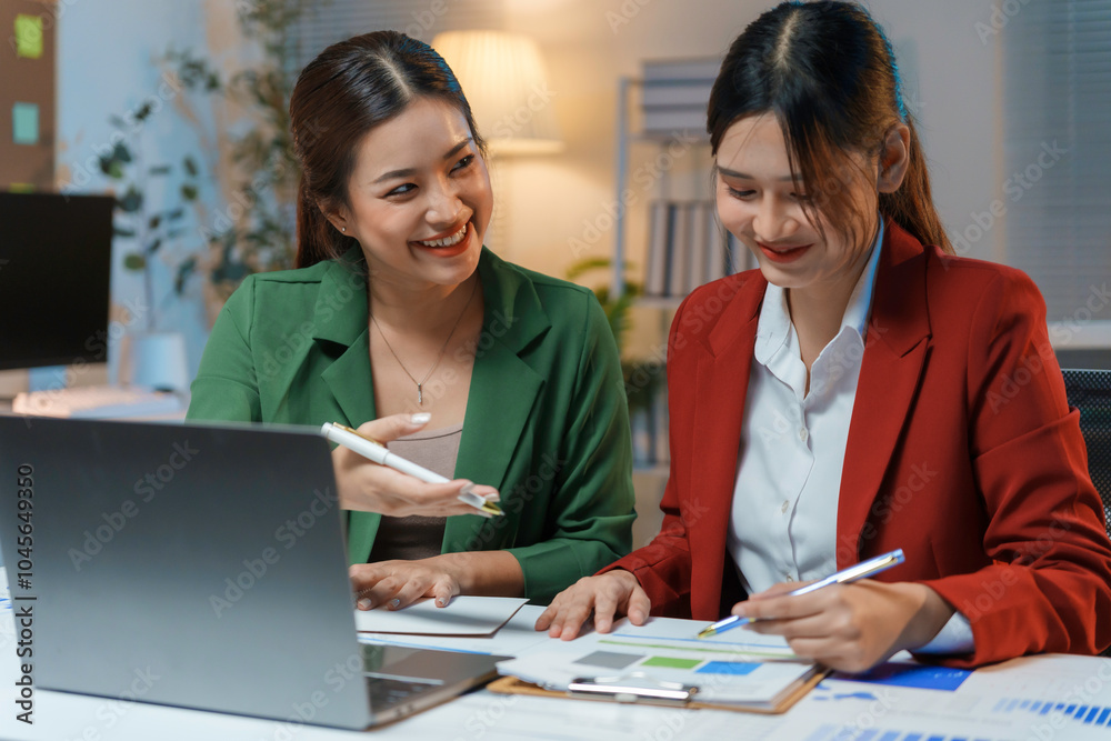 Two young businesswomen are working late in the office, collaborating on a project and analyzing financial data on a laptop Two young businesswomen are working late in the office, collaborating on a project and analyzing financial data on a laptop