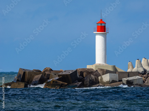 Small white lighthouse with red top and roof, concrete block breakwater of port.