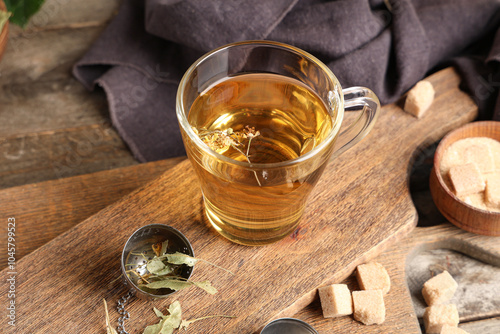 Billede på lærred Board with cup of tasty linden tea, sugar and metal infuser on wooden background