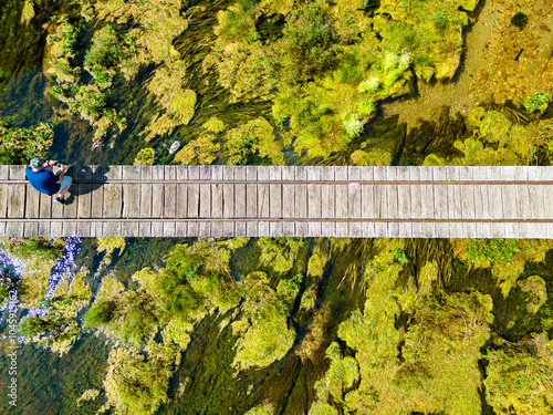 Aerial top down view of man kneeling on the wooden bridge crossing Gacka river in Majerovo Vrilo, Croatia, taking photos of ducks in the water photo