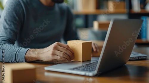 A man is working at a desk with a laptop and a cardboard box. He is holding the box in his hand and looking at the screen.