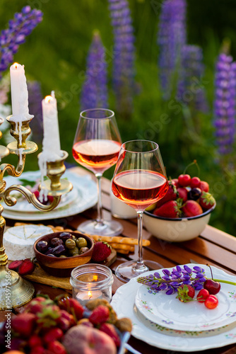 Beautiful elegant romantic dinner table decor, surprise day arrangement. Marriage proposal, engagement private party for two in a meadow. Sunset, golden hour. Cheese board, rose wine, fruits, lupine