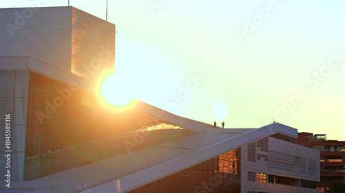 Dramatic sunrise view with silhouettes of people standing on the roof of the Oslo Opera House, capturing the sun’s warm glow and architectural details.