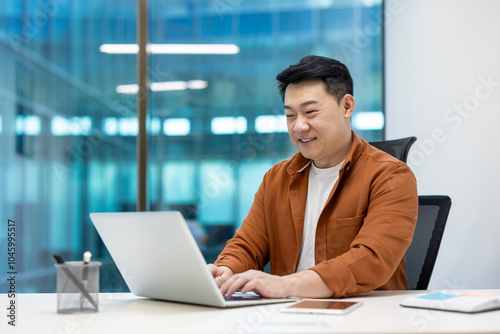 Asian businessman using laptop in bright modern office. Smiling while working, wearing casual shirt. Represents concepts of productivity, technology, teamwork, and professionalism. Includes phone.