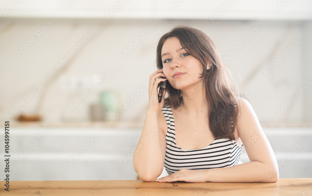 Fototapeta premium Young woman talking on mobile phone sitting at table in kitchen