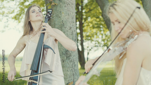 Two beautiful girls in dresses play violin and cello outdoors.
