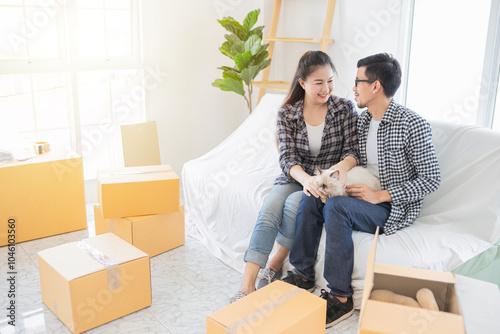 Happy couple asian lover takes a break from unpacking, sharing a moment on white couch surrounded by moving boxes. They carry Persian cat. cardboard boxes, playfully unpacking. Relocation concept.