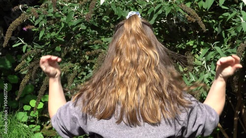 Woman showing her shiny natural golden blonde hair in the sun as a result of natural hair care treatments. Green tree in background. Beauty content. 