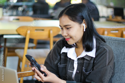 attractive and charming asian woman sitting in cafe using smartphone to listening music with earphone, browsing cyberspace and read news with laptop on desk, candid