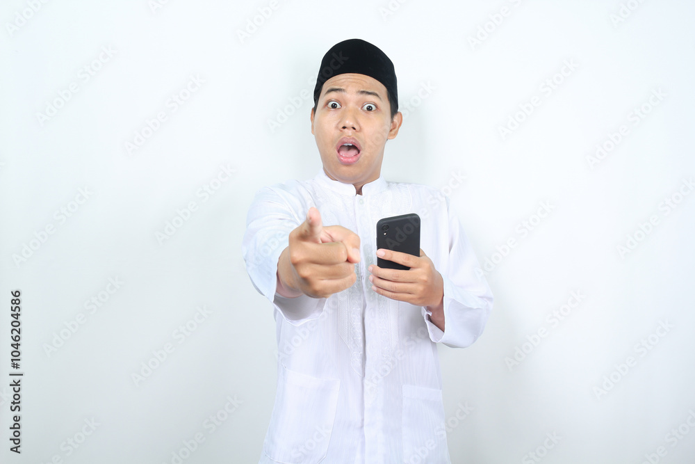 shocked asian muslim man pointing forward to camera with holding mobile phone shocked asian muslim man pointing forward to camera with holding mobile phone