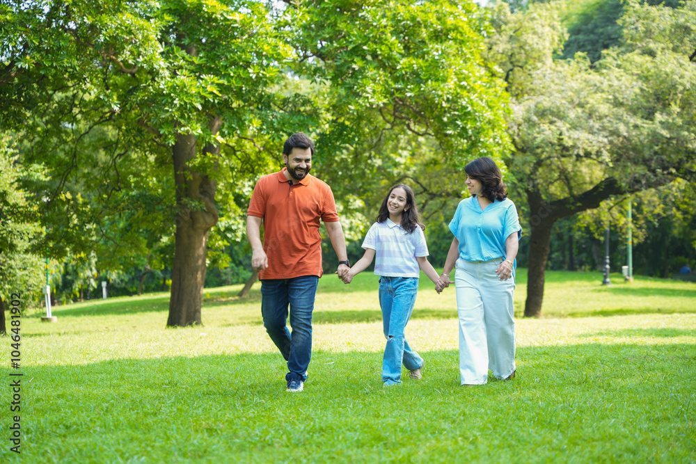 Obraz premium Happy young indian parents with child daughter holding hands and walking in summer park, playful family have fun together outdoor, Holidays and vacations, Kid bonding with mom and dad, togetherness.