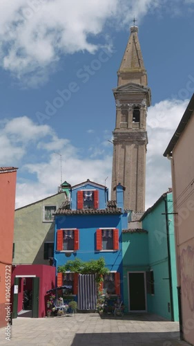 A bright blue house with red shutters stands under the iconic leaning bell tower in Burano, Venice, surrounded by colorful buildings under a partly cloudy sky.