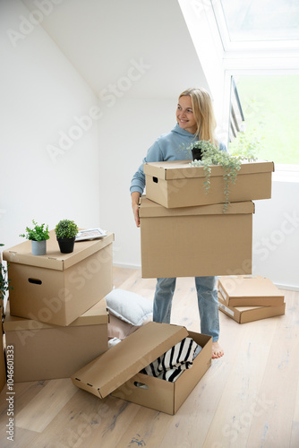 woman with blond hair stands in her new apartment, loft, house surrounded by many brown moving boxes and is carrying boxes